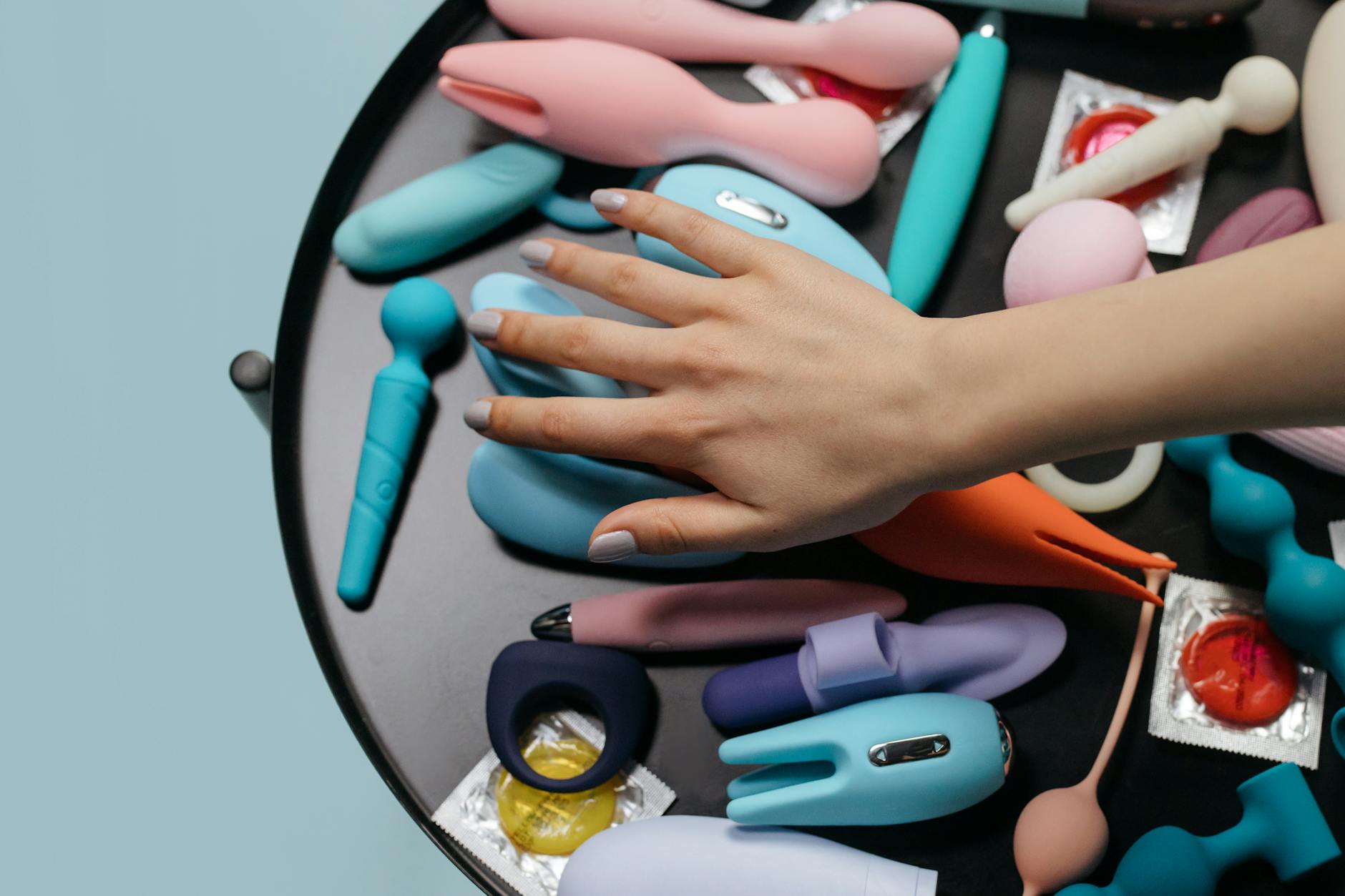 A hand reaching over a collection of colorful vibrators on a surface, symbolizing choice and personal agency in pleasure.